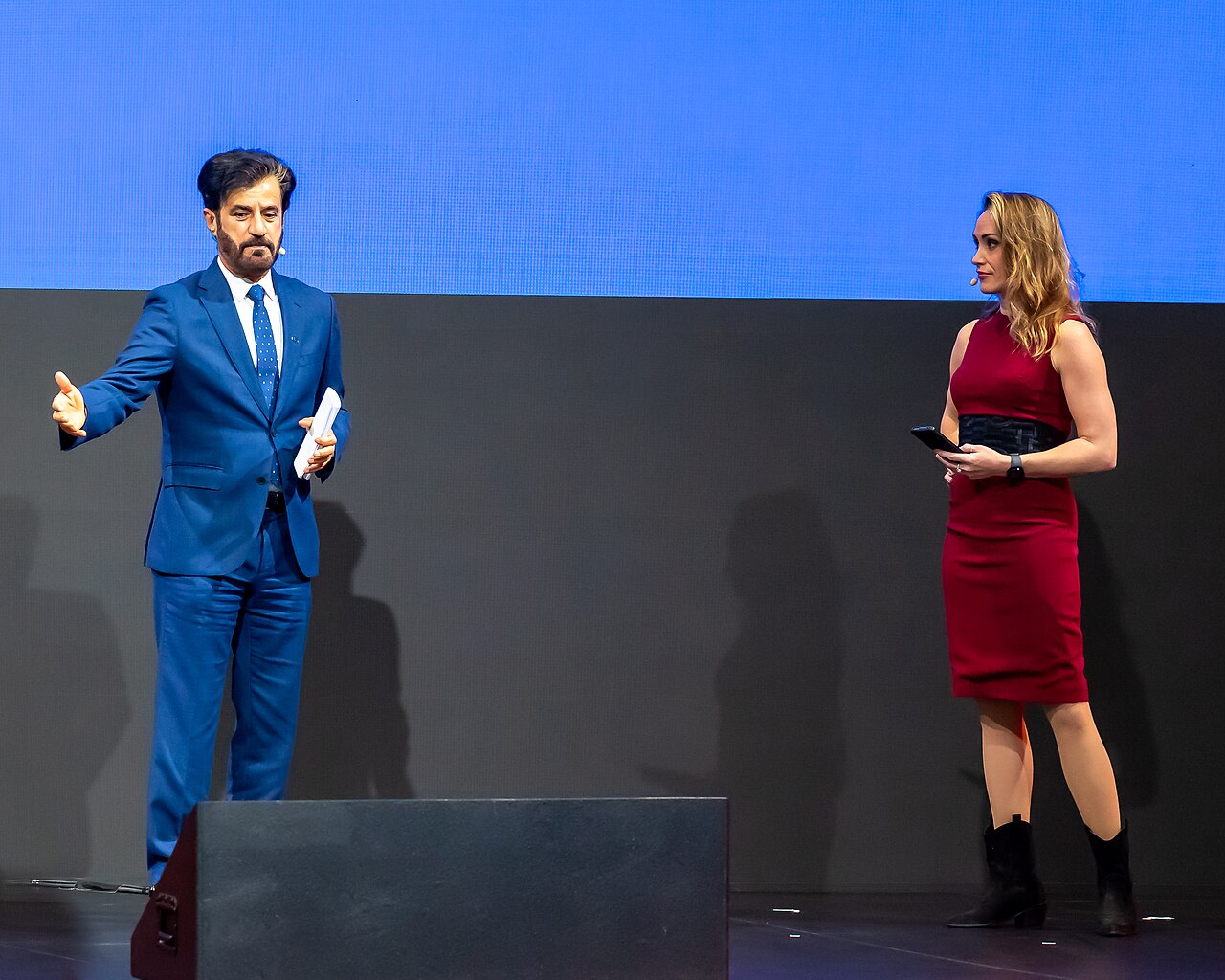 FIA president Mohammed Ben Sulayem in a blue suit speaking on stage, with a presenter in red beside him
