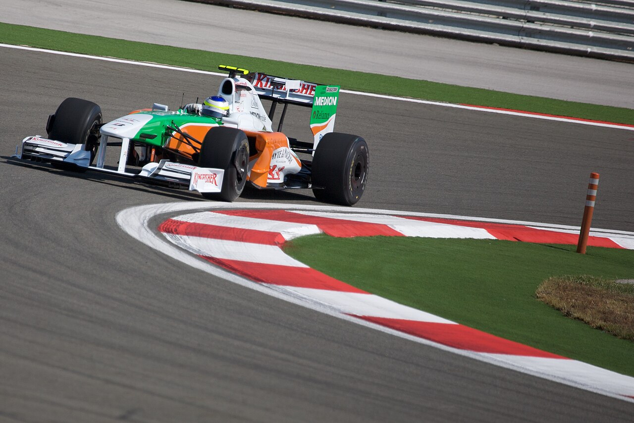 A white, green and orange Force India F1 car cornering over a red and white kerb