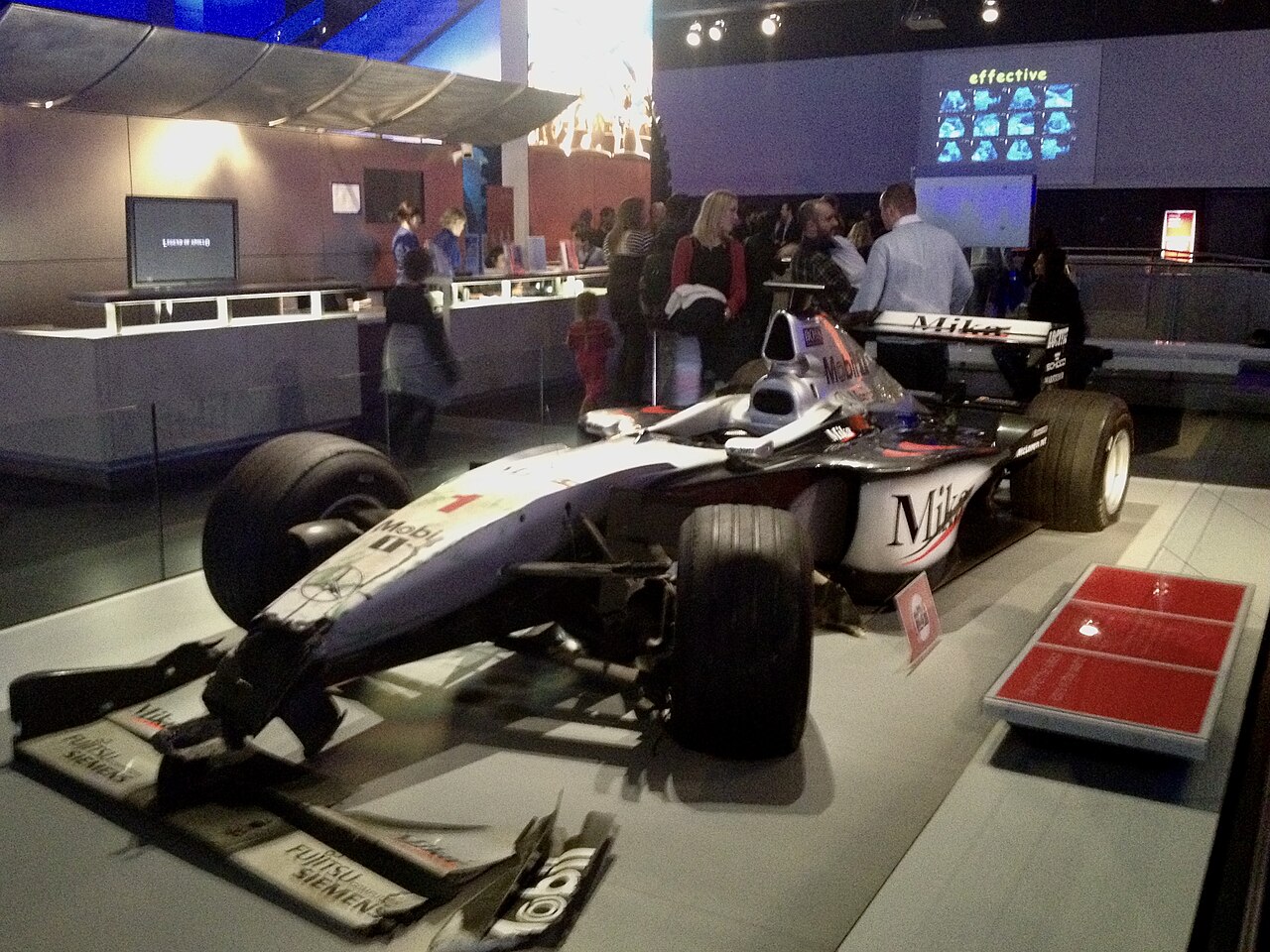 A silver and black McLaren F1 car with the number 1 on its nose, displayed in a museum gallery with visitors behind it