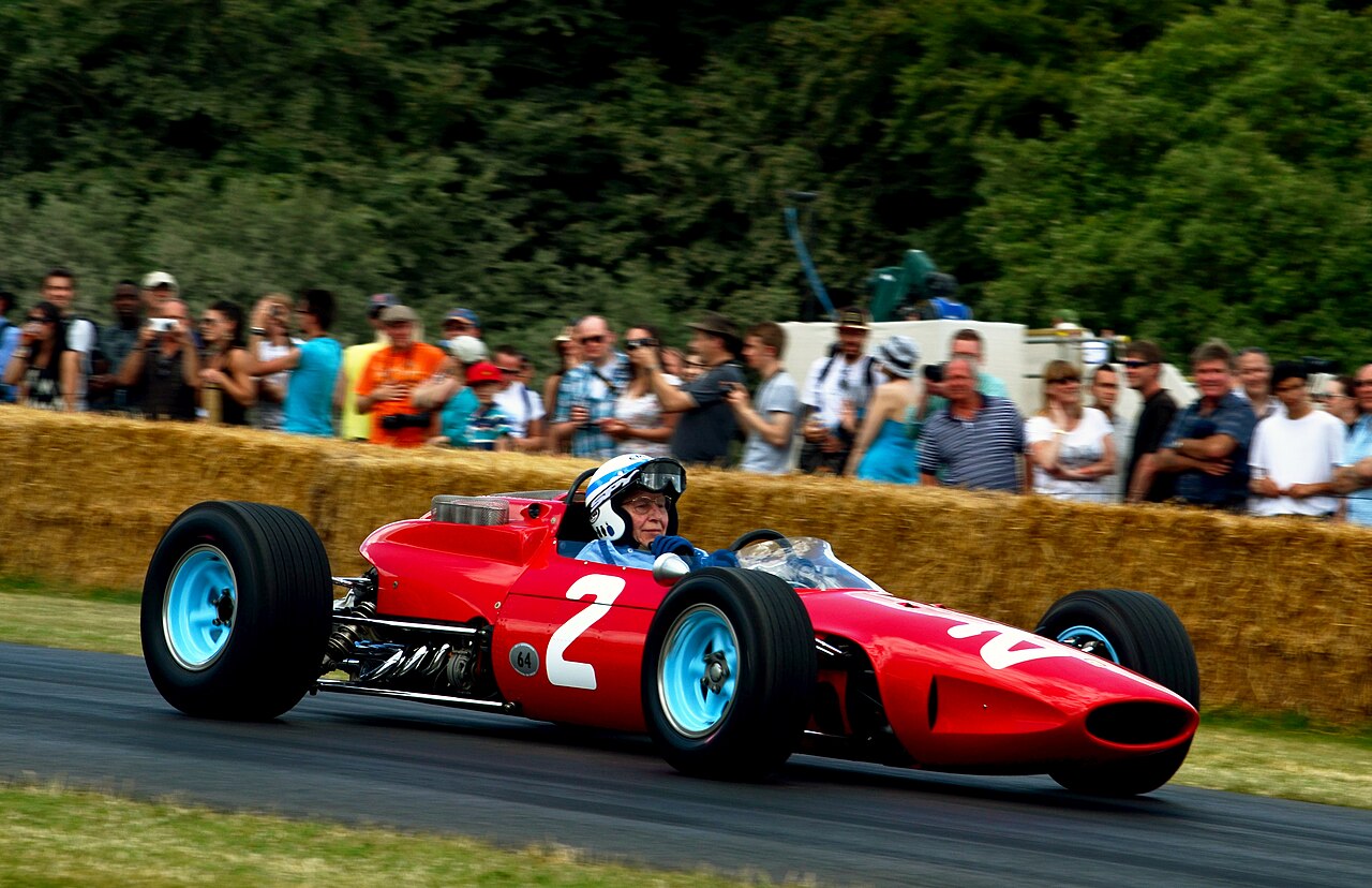 John Surtees, in a white helmet, driving a red Ferrari number 2 past a crowd behind straw bales